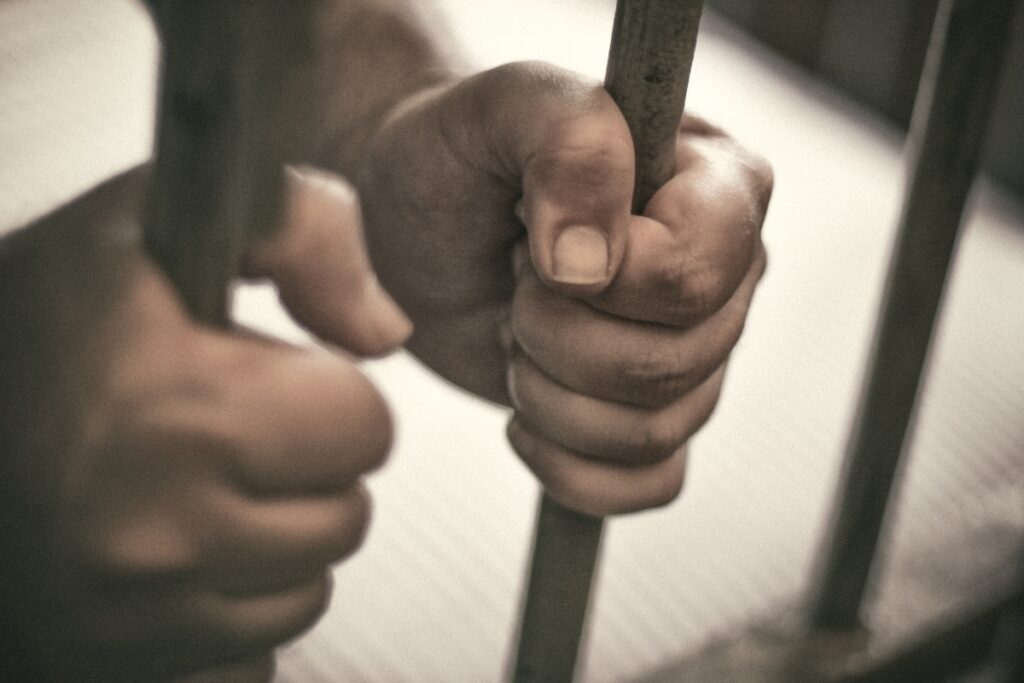 A man's hand holding barbed wire in a jail cell representing incarceration.