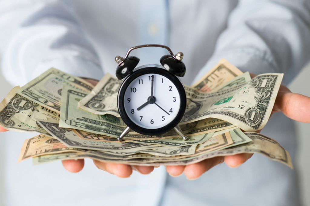 A person's hands holding money and an alarm clock representing the importance of time and cost savings.